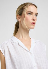 Blusa blanca de manga corta con cierre de botones en la parte delantera, cuello y una sutil textura de cuadros en una tela ligera.