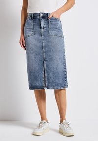 Jupe en denim, bleu clair, mi-longue avec fente frontale, deux poches avant et fermeture à bouton métallique. Associée à des baskets blanches.