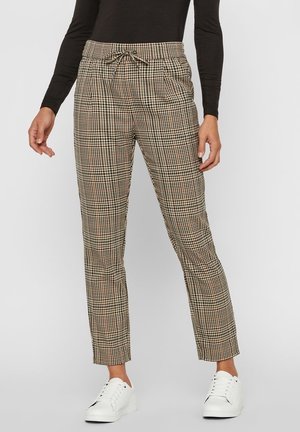 Femme portant un pantalon à carreaux beige et noir, longueur cheville, avec taille à cordon et baskets blanches, se tenant devant un fond uni.
