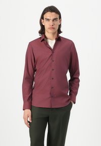 Chemise à manches longues pour homme, en tissu bordeaux profond, à boutons avec des boutons bleu marine, col structuré, portée avec un pantalon vert foncé.