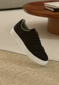 Fekete, alacsony szárú sportcipő fehér talppal bézs szőnyegen, egy kerek fából készült asztal közelében, amelyen egymásra rakott könyvek vannak.