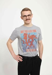 Grå bomullströja med färggrafisk tryck av två tecknade karaktärer med text, tillsammans med svarta byxor och ett svart bälte.