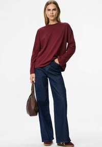 Ciemnoczerwona koszula z długim rękawem, luźna, w połączeniu z ciemnoniebieskimi spodniami dżinsowymi o szerokich nogawkach. Brązowa torba w ręku, minimalna biżuteria.