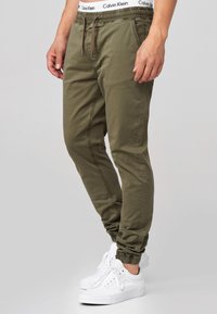 Olive green jogger pants with elastic waistband, adjustable drawstring, and cuffed ankles, paired with white sneakers.