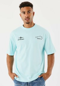 Jeune homme avec des cheveux bouclés courts et des boucles d'oreilles portant un t-shirt bleu clair "Project X Paris" et un jean bleu avec du texte imprimé, les mains dans les poches.