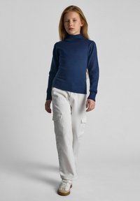 Mornarska turtleneck pletena jopica v kombinaciji z belimi cargo hlačami. Hlače imajo stranske žepe. Model nosi bele superge s svetlo rjavimi detajli.