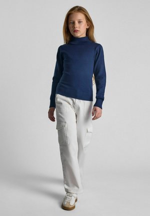 Navyfarbener Rollkragenpullover, kombiniert mit weißen Cargo-Hosen. Die Hosen verfügen über Seitentaschen. Das Model trägt weiße Sneakers mit beigen Akzenten.