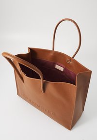 Brown leather tote bag with textured surface, two handle loops, and purple interior lining. Features a brand tag inside.