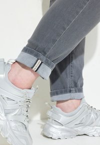 Jeans de mezclilla gris con puños enrollados que revelan una etiqueta, combinados con zapatillas deportivas plateadas de diseño de materiales mixtos y acentos texturizados.
