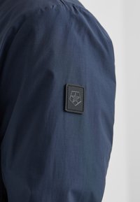 Marineblaue Jackenstoff mit glatter Textur, versehen mit einem schwarzen rechteckigen Logo-Patch am linken Ärmel. Einfaches und modernes Design.