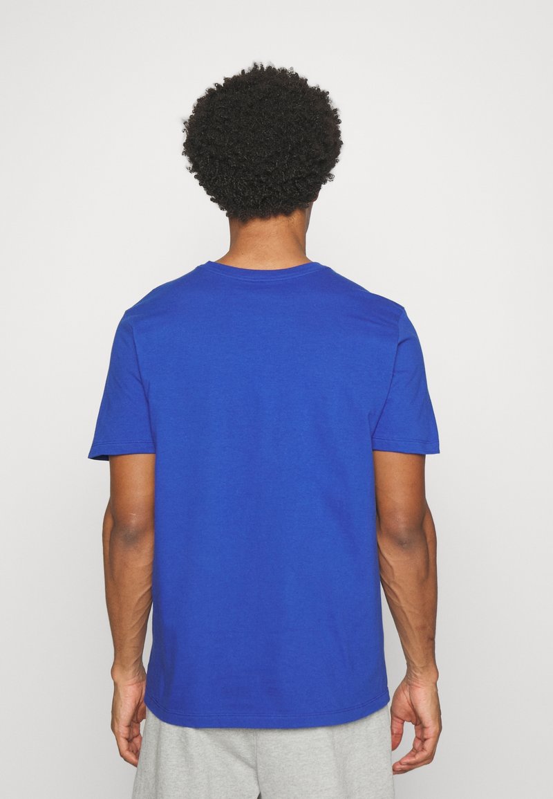 Homme aux cheveux bouclés portant un t-shirt bleu uni à manches courtes et un pantalon gris clair, de dos sur fond blanc.