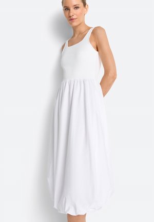 Donna che indossa un abito midi bianco senza maniche con corpetto aderente e orlo a palloncino raccolto, in piedi su sfondo bianco.