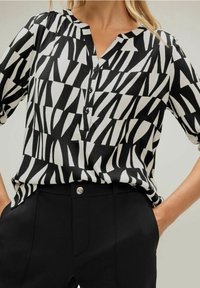 Blusa motivo a righe bianco e nero con scollatura a V, maniche a tre quarti e pattina dei bottoni. Abbinata a pantaloni neri su misura.