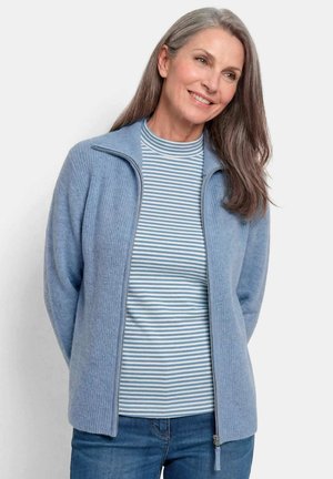 Donna sorridente con capelli lunghi e grigi che indossa un maglione blu con zip sopra una camicia a righe blu e bianche e jeans blu su sfondo bianco.