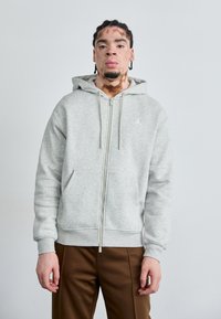 Grå zip-up hoodie med en kængurulomme, snoretræk i hætten og kontrasterende logo. Stylized med brune bukser, tekstureret stoffinish.
