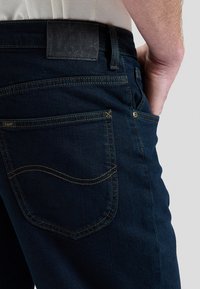 Asmuo dėvi tamsiai mėlynas Lee džinsus su matoma galine kišene ir prekės ženklo etikete, ranka ilsisi priekinėje kišenėje, šviesi fonas.