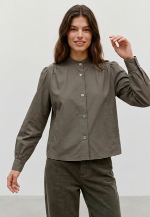 Femme aux cheveux bruns ondulés portant un chemisier vert olive boutonné et un pantalon en velours côtelé, souriante avec une main levée.