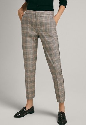 Person iført slim ternede bukser med hænderne i lommerne og sorte loafers, der står mod en neutral baggrund.