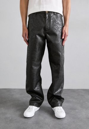Black faux leather pants with a textured floral pattern, relaxed fit, and a gold button at the waistband, paired with white sneakers.