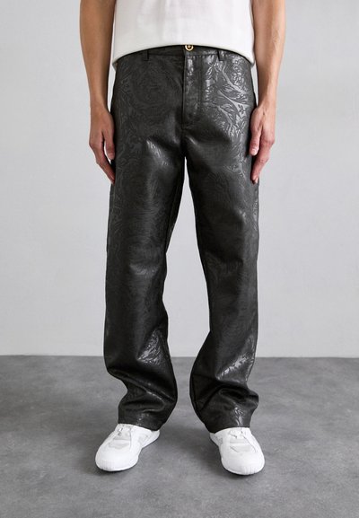 Pantalons en simili cuir noir avec un motif floral texturé, coupe décontractée et un bouton doré à la taille, associés à des baskets blanches.