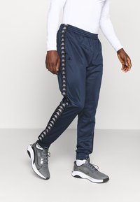 Pantalones atléticos azul marino con cintura elástica, franjas laterales con logo y piernas ajustadas, combinados con zapatillas deportivas grises.