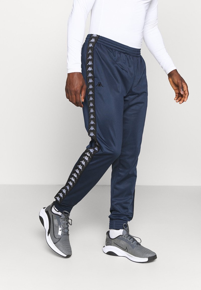 Pantalones atléticos azul marino con cintura elástica, franjas laterales con logo y piernas ajustadas, combinados con zapatillas deportivas grises.