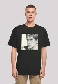 Sort t-shirt med et firkantet grafisk print af en blyantsketch af en mands ansigt, detaljeret udtryk og tekstureret hår, centreret på brystet.