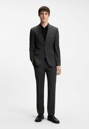 Młody mężczyzna ubrany w ciemnoszary, dopasowany garnitur i czarną koszulę, stojący z jedną ręką w kieszeni na jednolitym białym tle.