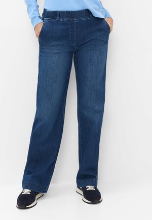 Person iført mørkeblå jeans med lige ben og elastisk talje, afslappede sorte sneakers og lyseblå langærmet top med hænderne i lommerne.