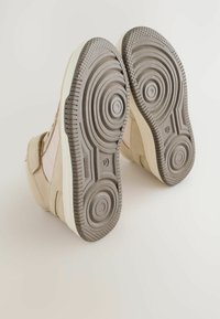 Baskets beiges avec une tige texturée et un motif de semelle circulaire sur la semelle en caoutchouc grise. Détails de bande Velcro et accents ronds visibles.