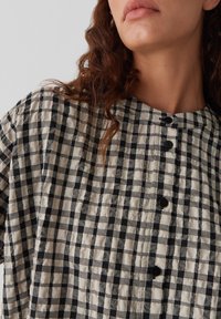 Camicia a quadri in bianco e nero con superficie testurizzata, caratterizzata da scollatura rotonda, cuciture verticali e bottoni neri sul davanti.
