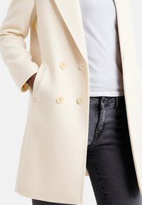 Cappotto crema doppiopetto con due grandi bottoni, tasche laterali e una silhouette dritta, abbinato a una camicia bianca e jeans scuri.