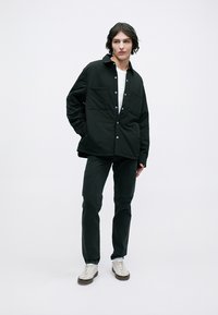 Schwarze Knopfjacke mit zwei Fronttaschen, kombiniert mit dunklen Jeans und weißen Sneakers. Der Stoff wirkt texturiert und strapazierfähig.