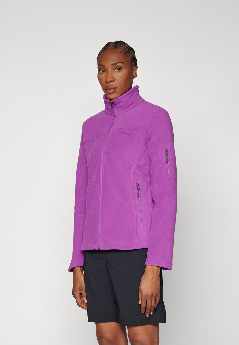 Giacca in fleece viola con colletto alto, dotata di tasche con zip e una texture liscia. Indossata sopra pantaloni corti neri.