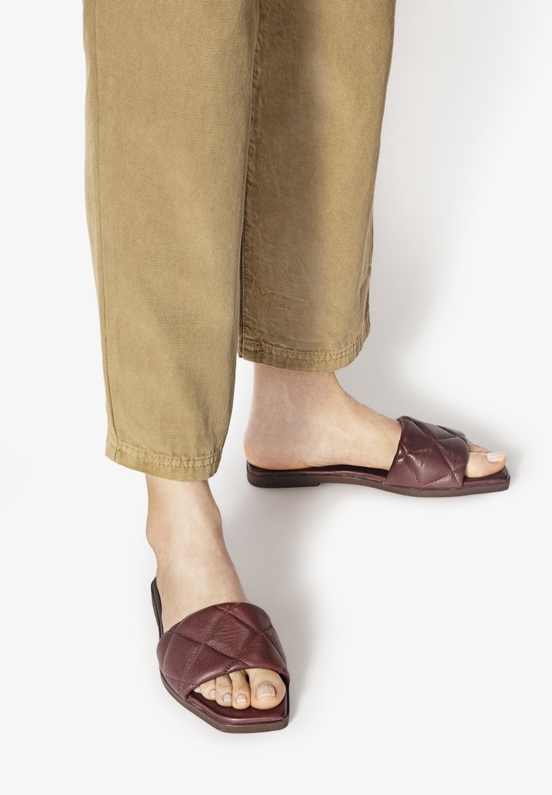 Pieds portant des sandales marron matelassées à enfiler, associées à un pantalon court marron clair sur fond blanc.