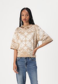 Beige kortermet topp med hvite paisley-mønstre og rund hals. Kombinert med blå jeans. Materialet ser mykt ut med en avslappet passform.
