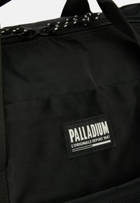 Saco de tecido preto com superfície texturizada, com fecho de zip e uma alça trançada. Inclui um emblema branco com o texto "PALLADIUM".
