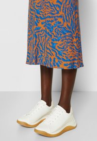 Baskets blanches au design texturé, avec une semelle en caoutchouc et une fermeture à lacets, associées à une jupe à motifs orange et bleu.