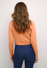 Mujer con largo cabello ondulado castaño, vistiendo un crop top naranja de mangas largas y unos vaqueros de talle alto azules, vista desde atrás.