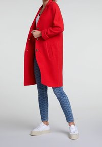 Persona che indossa un lungo cappotto rosso brillante, jeans blu con fantasia, scarpe da ginnastica bianche e una maglietta bianca, in piedi contro uno sfondo semplice.