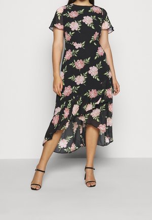 Vrouw draagt een zwarte high-low jurk met bloemenprint van roze bloemen en groene bladeren, gecombineerd met zwarte enkellaarsjes met enkelbandje.