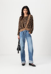 Cardigan marron à imprimé léopard avec un devant à boutons, associé à un jean taille haute bleu clair et des chaussures noires à enfiler. Sac à main noir tenu à la main.