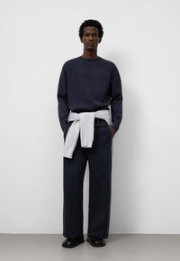 Mörkblå stickad tröja med rundad halsringning, kombinerad med vida gråbyxor och en grå tröja knuten i midjan; svarta skor fulländar outfiten.