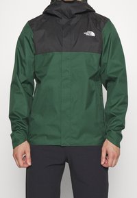 Veste de pluie à capuche verte et noire avec une finition résistante à l'eau, dotée d'une fermeture éclair frontale, de poignets élastiques et d'un logo sur la poitrine.