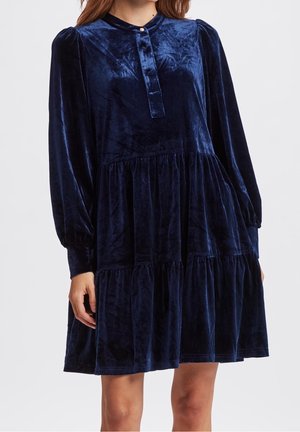 Kobieta ubrana w długą, granatową welurową sukienkę z marszczonymi warstwami i zapinanym kołnierzykiem, stojąca na jednolitym tle.