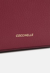 Burgundská kožená taška s texturovaným povrchem, zlatým logem "Coccinelle" a čistými liniemi s minimalistickým designem.