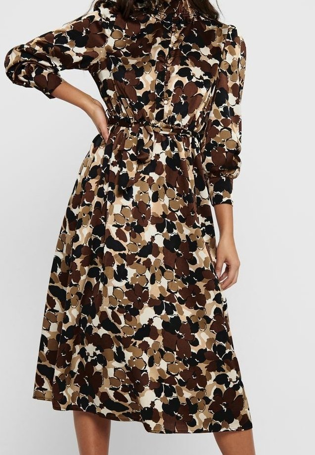 Femme portant une robe longue jusqu'aux genoux, à manches longues, avec un motif floral abstrait marron, noir et beige, et une taille cintrée.