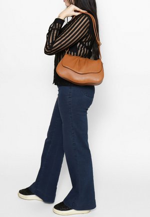 Brown leather shoulder bag with a curved design, featuring a smooth texture and a single strap, carried by a person wearing dark jeans.