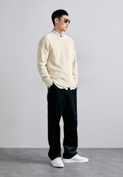 Beige ribbed sweater over a light blue shirt, paired with dark trousers and white sneakers. Model wears aviator sunglasses.