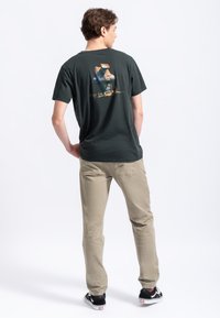 Donkergroen katoenen t-shirt met een achterafbeelding van een figuur op een machine; gecombineerd met licht kaki broek en zwarte sneakers met witte accenten.